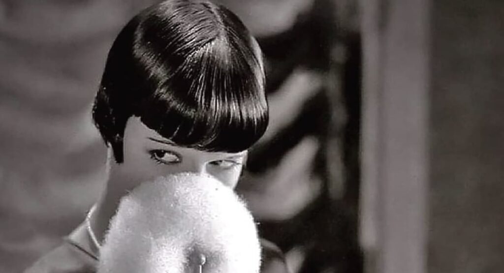 Film still from Love ’Em and Leave ’Em — Louise Brooks peering over a white fur muff that covers the lower half of her face, glossy black bob, eyes cut sharply to the side