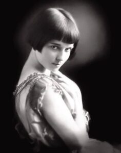 Studio portrait of a teenaged Louise Brooks looking back over a bare shoulder, dark hair in an early bob, wearing an embroidered sleeveless dress against a black background