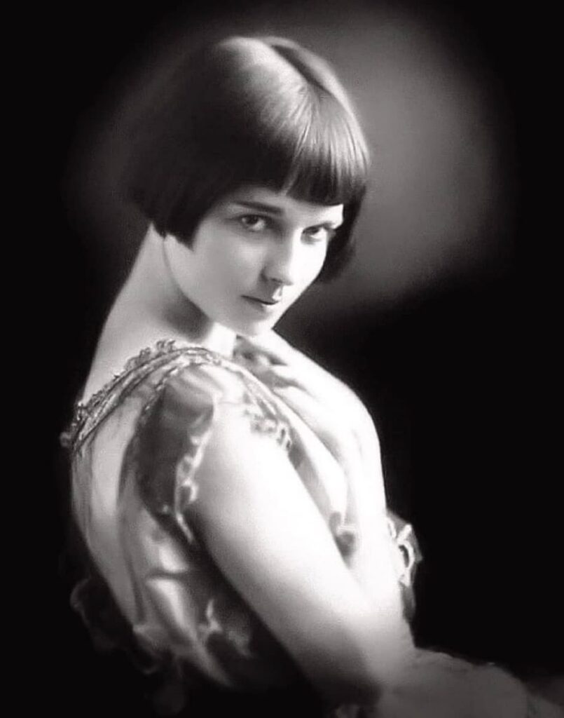 Studio portrait of a teenaged Louise Brooks looking back over a bare shoulder, dark hair in an early bob, wearing an embroidered sleeveless dress against a black background