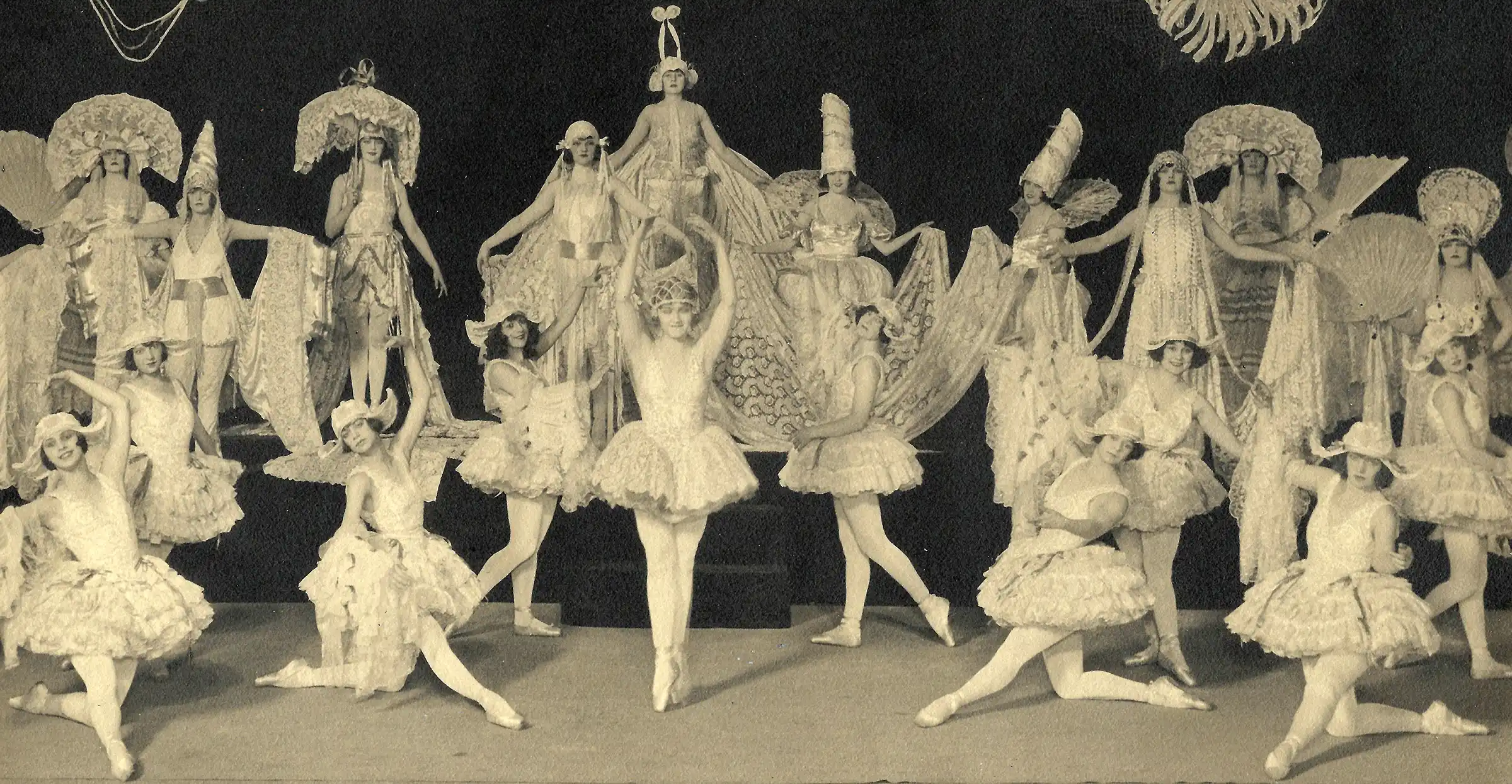 Nineteen dancers in elaborate lace tutus and towering lace headdresses pose on a Broadway stage; at center, Mary Eaton stands en pointe with her arms raised overhead.