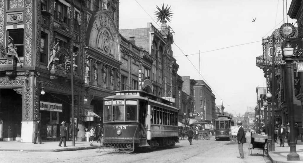 Baltimore street scene about 1909 — streetcar number 1533 passing an ornately carved theater and museum front, men in straw hats on the sidewalk, and a horse-drawn wagon further up the block
