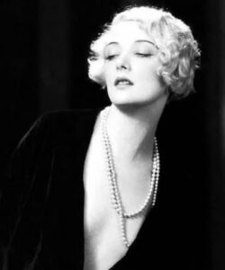 Glamour portrait of Mary Nolan, blond and wearing a long strand of pearls with her eyes closed, against a dark background.