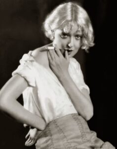 Studio portrait of Mary Nolan in a white dress, one hand on her hip, glancing over her shoulder.