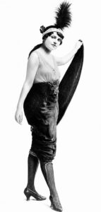 Full-length photograph of a young woman in a plumed headband, sleeveless bodice and dark hobble skirt, holding a long cloak out to one side, reproduced in the 1915 newspaper feature as the improved 1915-model Show Girl.
