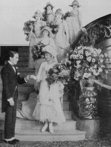 Wedding scene from the 1918 film His Majesty, Bunker Bean, with bridesmaids in wide-brimmed hats gathered on a staircase and a man in formal dress at lower left. Public domain.