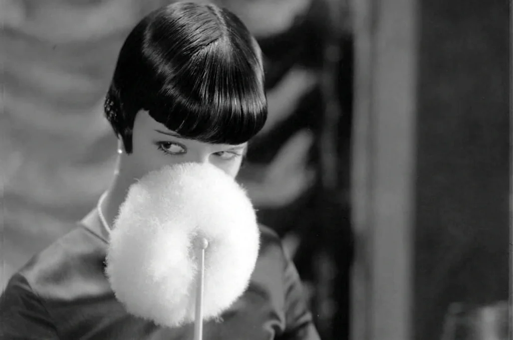 Louise Brooks in Love ’Em and Leave ’Em (1926), peering over a large white powder puff that hides the lower half of her face, her black bob catching the studio light