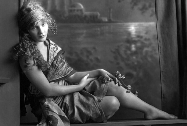 Mary Miles Minter in a studio portrait, seated barefoot on a ledge holding a sprig of blossoms, a painted lakeside backdrop behind her.