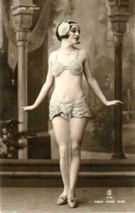 Postcard of a young Lupe Velez in a beaded two-piece stage costume and feathered headband, posed before a painted backdrop