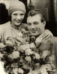 Lupe Velez in a cloche hat smiling beside Gary Cooper, with a bouquet of roses in front of them