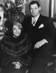 Lupe Velez in a fur coat and small tilted hat, seated beside Johnny Weissmuller in a dark suit