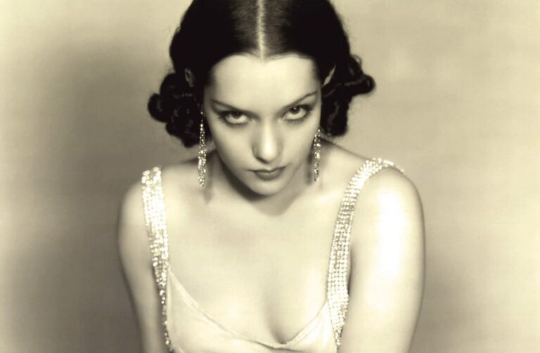 Lupe Vélez in a 1930s studio portrait, dark curls and drop earrings, glaring at the camera in a satin gown with rhinestone straps.