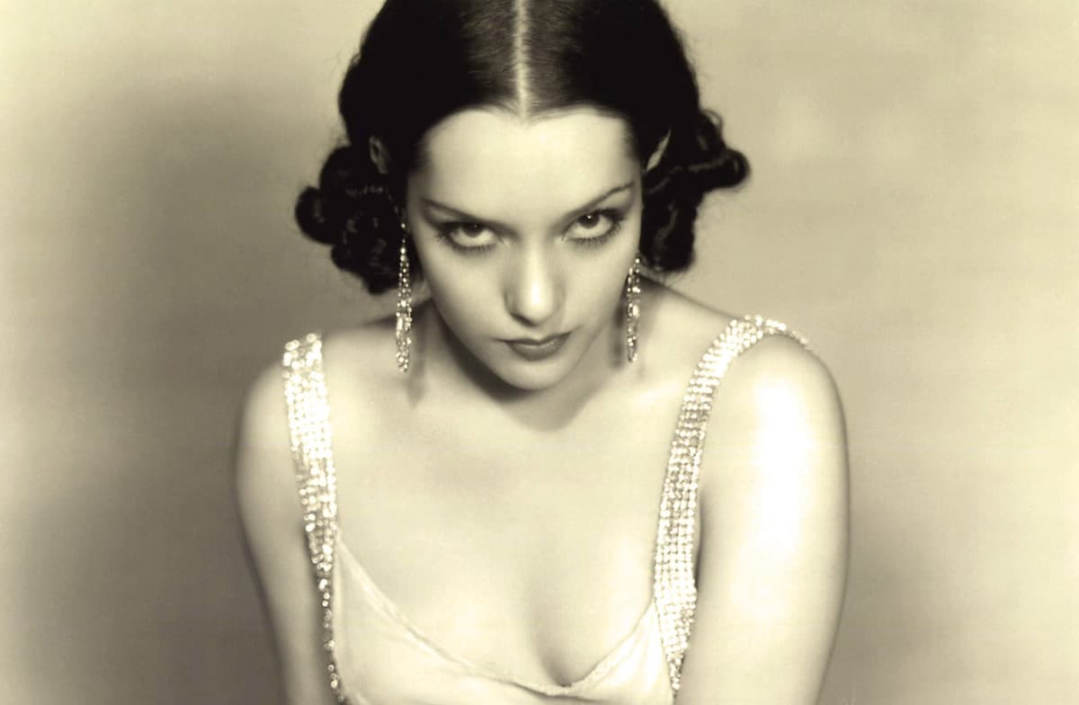 Lupe Vélez in a 1930s studio portrait, dark curls and drop earrings, glaring at the camera in a satin gown with rhinestone straps.